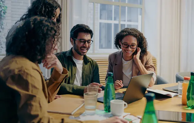 A group of professionals collaborate around a conference table in a bright office, reviewing information on laptops and discussing work.