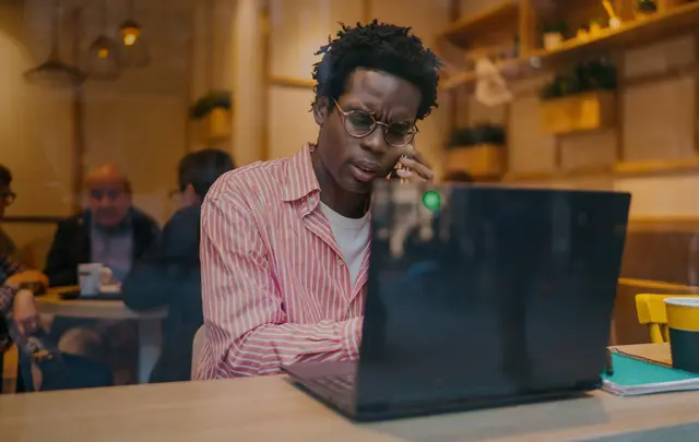 A professional works on a laptop while speaking on a phone in a café or coworking environment.