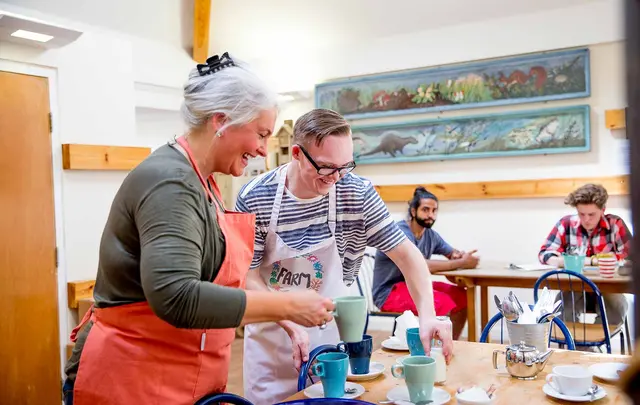a young man and an older woman in aprons placing drinks on a cafe table