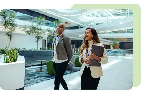 Two professionals wearing visitor badges walk through a bright, modern office atrium with curved architectural features, indoor trees, and collaborative seating areas.