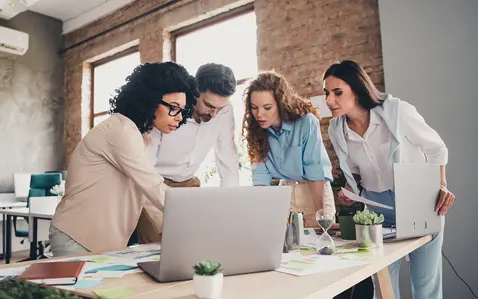 Four professional working on a laptop at a desk in a modern office.
