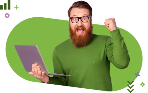 A joyful man with a beard and glasses raises his fist while holding a laptop. 