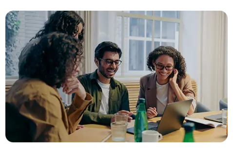 A group of professionals collaborate around a conference table in a bright office, reviewing information on laptops and discussing work.