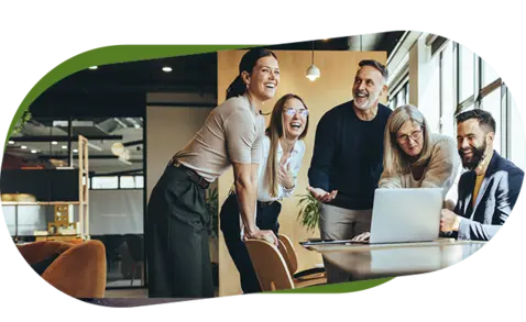 Five professionals gathered around a table in a modern office, smiling and engaged as they look at a laptop. 