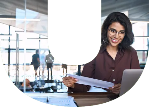 A woman wearing glasses and a dark brown shirt sits at a desk, holding documents in her left hand while focused on a laptop in front of her.