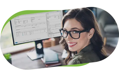 A smiling woman with black-rimmed glasses sits at a wooden desk, looking over her shoulder at the camera.