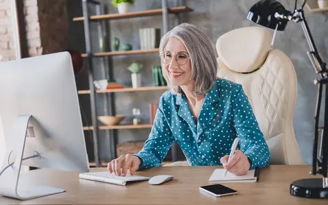 A professional woman sits at a desk in a modern home office, working on a desktop computer while taking notes in a notebook, with shelves, plants, and a desk lamp visible in the background.