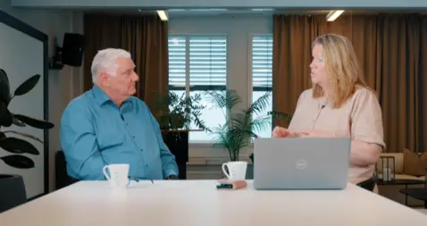 Two people seated at a table with a laptop and mugs, engaged in a discussion in a meeting room setting.