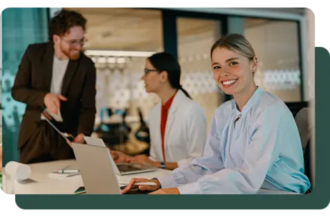 Three people working at laptops in a modern office, with one person in the foreground seated at a desk and two colleagues collaborating in the background.