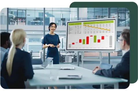 A presenter standing beside a screen displaying bar charts and financial data during a meeting with colleagues in a modern office.