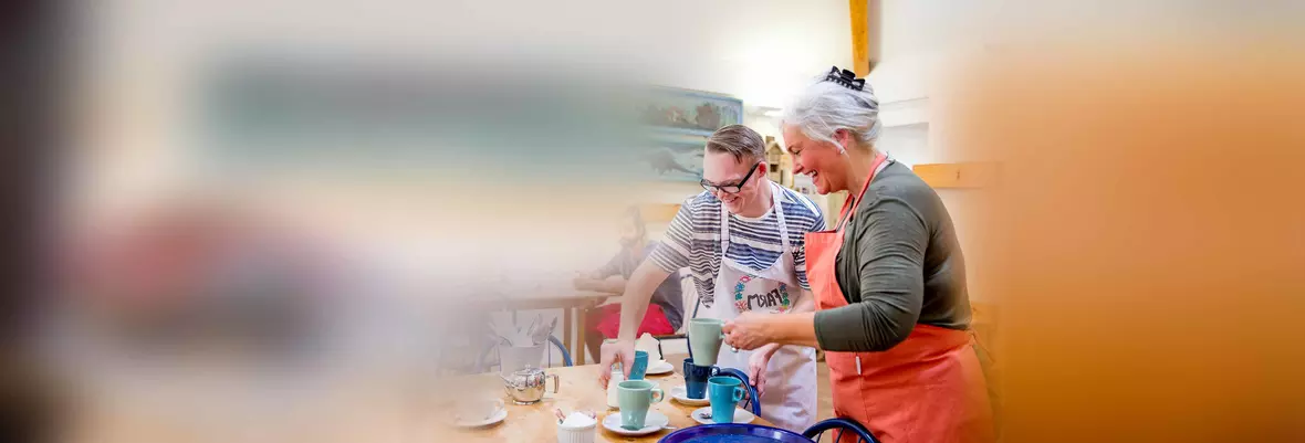 a young man and an older woman in aprons placing drinks on a cafe table