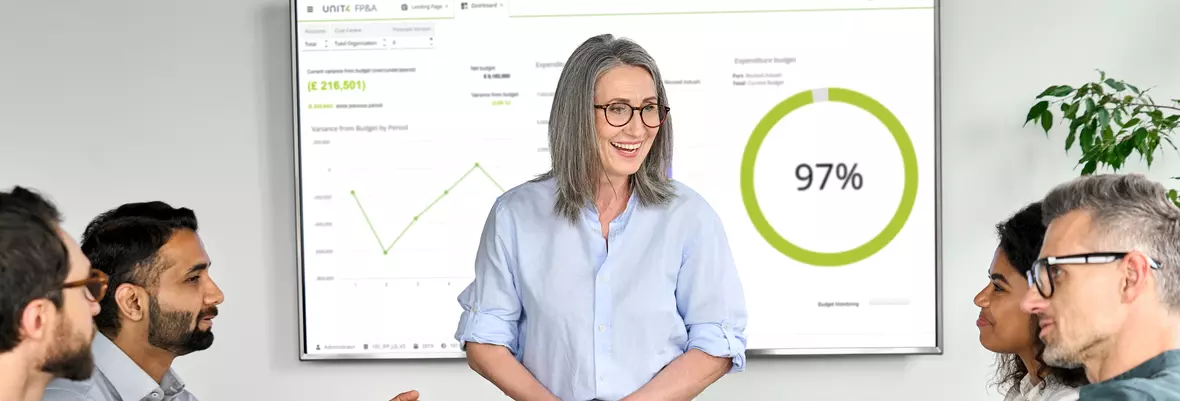 Smiling woman talking to people around a table. Behind her a screen with Unit4 FPA