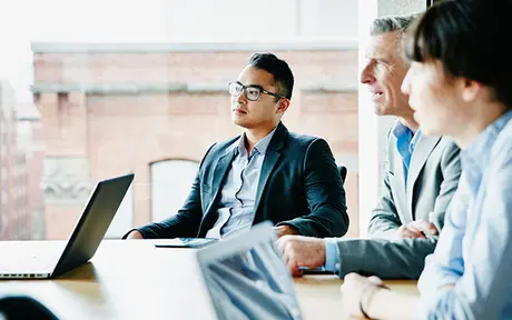 Three professionals sit around a wooden conference table in a modern office with large windows and exposed brick walls.