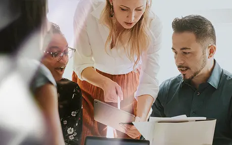 A group of three people engaged in a discussion, one showing a tablet to others. 