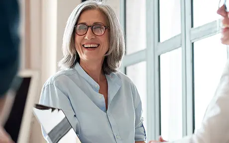 Two people are seated near large windows in a bright indoor setting, engaged in conversation.