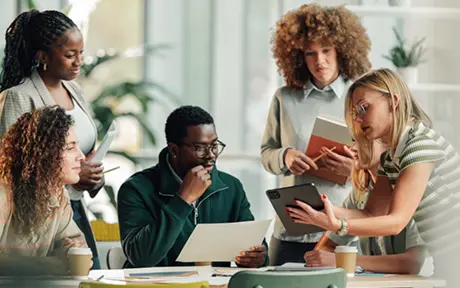 A diverse group of professionals collaborating around a table in a modern office, reviewing documents, a laptop, and a smartphone, with notebooks and coffee cups visible, illustrating teamwork, data analysis, and business decision-making.