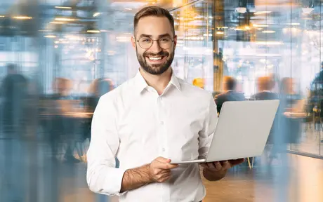 Smiling professional holding a laptop in a modern office environment with blurred colleagues working in the background