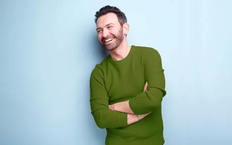 a smiling man in a green jumper leaning on a blue background