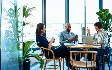 three people talking around a circular table which has their laptops resting on it