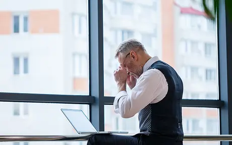 A person in business attire sits at a desk with a laptop, appearing stressed or thoughtful, in front of a large window with a cityscape view.