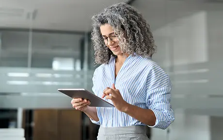 Person mit lockigem grauem Haar, Brille und gestreiftem Hemd, lächelt, während sie in einer modernen Büroumgebung ein Tablet verwendet.