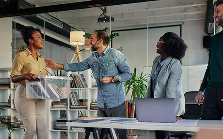 Two professionals shaking hands in an office setting with two other colleagues watching and smiling.