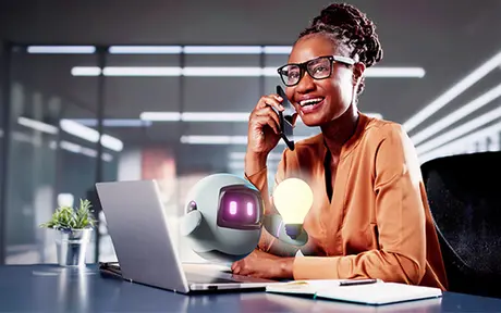 Henkilö istuu toimistossa työpöydän ääressä, puhuu puhelimeen ja käyttää kannettavaa tietokonetta, ja hänen vierellään on pieni robotti.