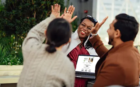 Three people engage in a cheerful conversation outdoors, with one person high-fiving another while the third person holds an open laptop.