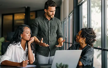 Three professionals in a discussion around a laptop in a modern office setting.