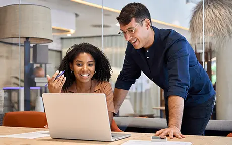 Two people are smiling and looking at a laptop in a modern office setting. They are sitting and standing at a table with papers. The environment is bright with a large window and contemporary decor.