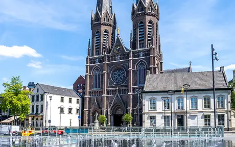 Stadsplein in Tilburg met fontein en de Heuvelse Kerk op de achtergrond, omringd door historische gebouwen en groen onder een blauwe lucht.