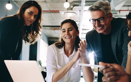 A group of people gathered around a laptop, appearing engaged and cheerful in a modern office setting. One person is holding a pen, and another is clapping. They are working collaboratively.