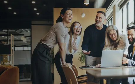 A group of people joyfully collaborating around a laptop in a modern office setting with large windows and indoor plants.